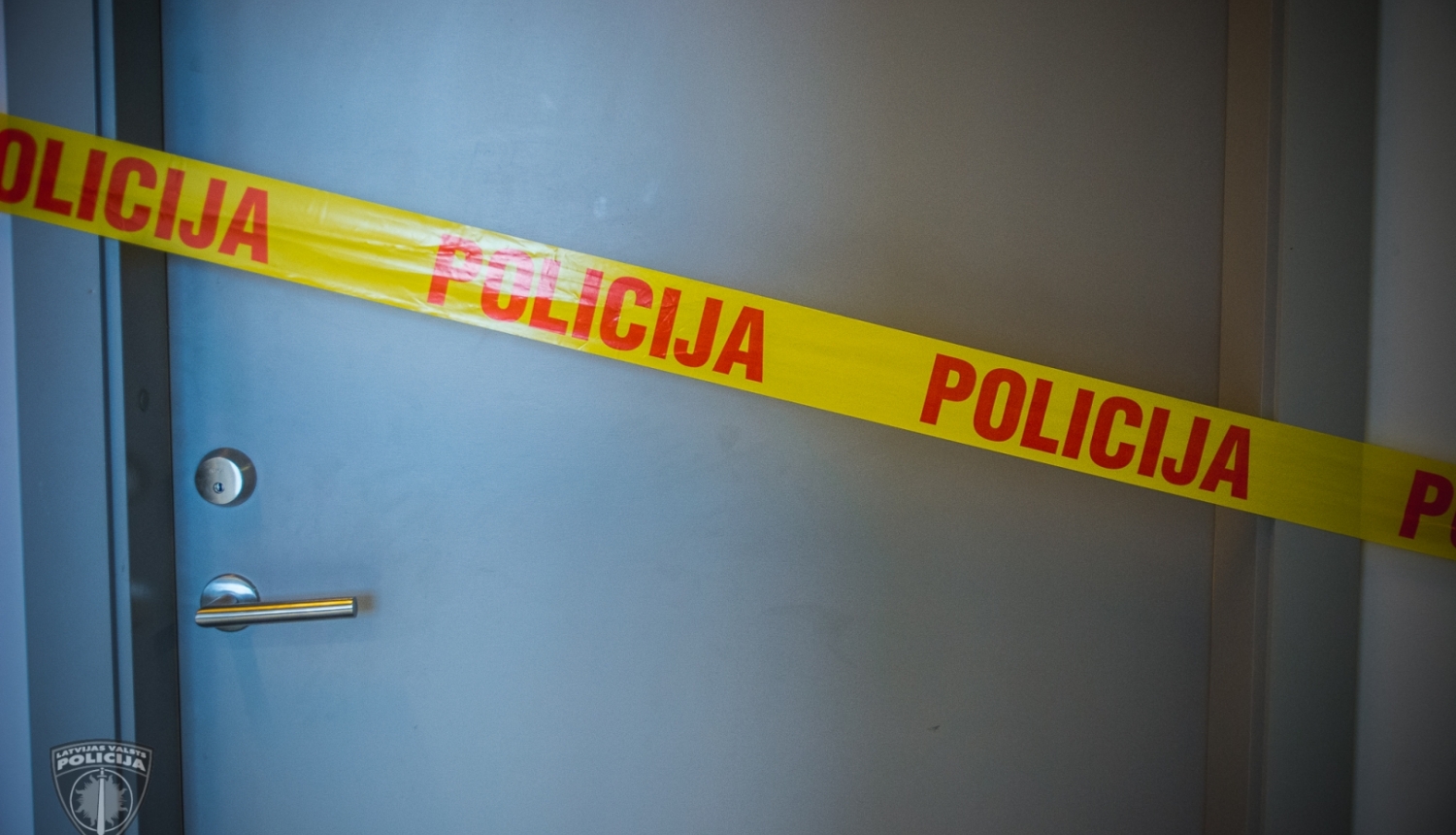 Aizvērtas durvis, kurām priekšā pa diagonāli novilkta dzeltena lenta, uz tās ar sarkaniem burtiem uzrakstīts POLICIJA